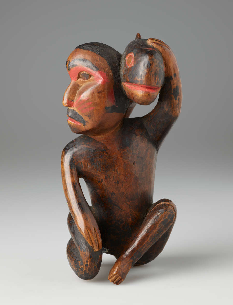 Side-view of worn painted wooden sculpture of a kneeling man looking aside, holding a small whale against his head. The man's clothes and hair are rendered in painted black forms.