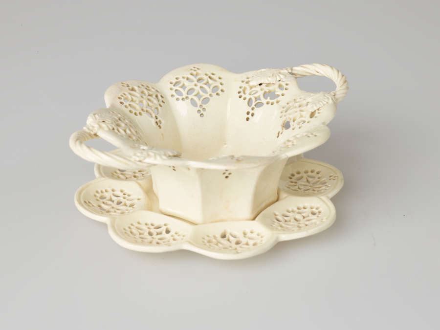 A cream-colored dish with two swirled handles and pierced decorations. Dish sits on a matching tray.