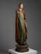 A side view of an aged wooden statue, draped in faded green and red garments standing on a carved pedestal.