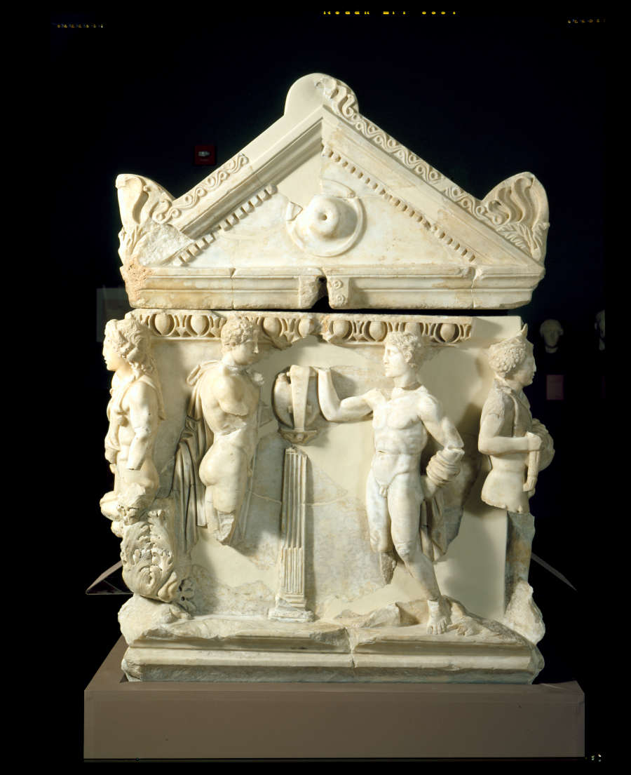 A panel of a marble coffin with a triangular roof and deep set carvings of standing figures engaging in conversation. 