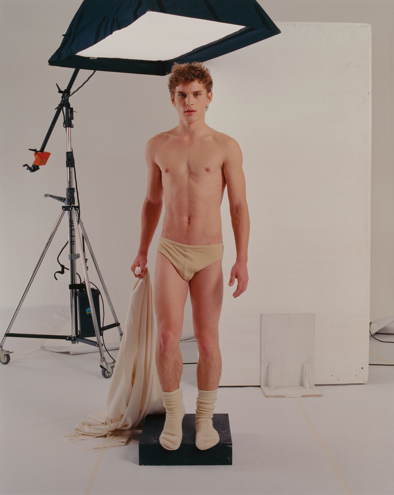 A fair-skinned male figure in beige underwear and socks stands on a black platform, holding draped fabric, and gazes into the camera in a studio setup with professional photography equipment. 