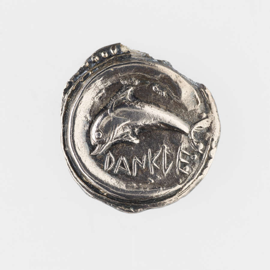 Silver coin with irregular edges. Embossed on its surface is a fish soaring, below which is Greek lettering.