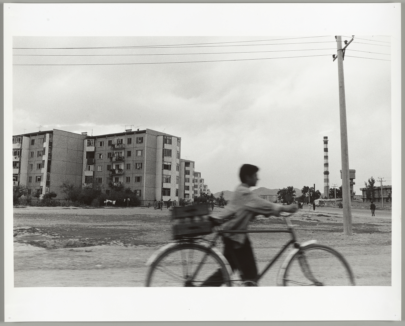 A black-and-white scene of a blurred cyclist in the foreground rides past a desolate landscape of block-style apartments and a smokestack with power lines stretching across the sky. 