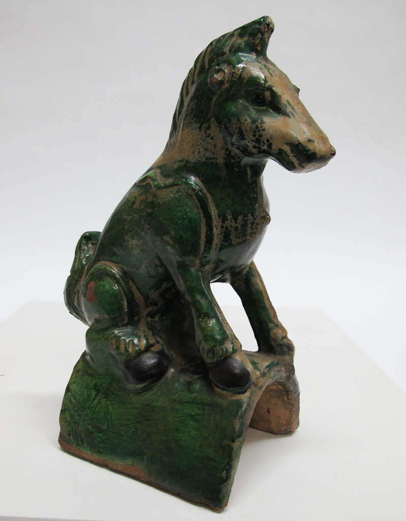 Side view of a green and brown glossy sculpture of a seated horse on an arched platform. The horse’s face and side of its necks feature larger patches of brown. 