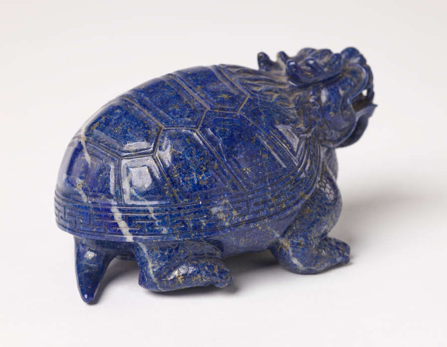 A back view of a blue sculpture of a horned tortoise, whose surface is covered with carved geometric patterns.