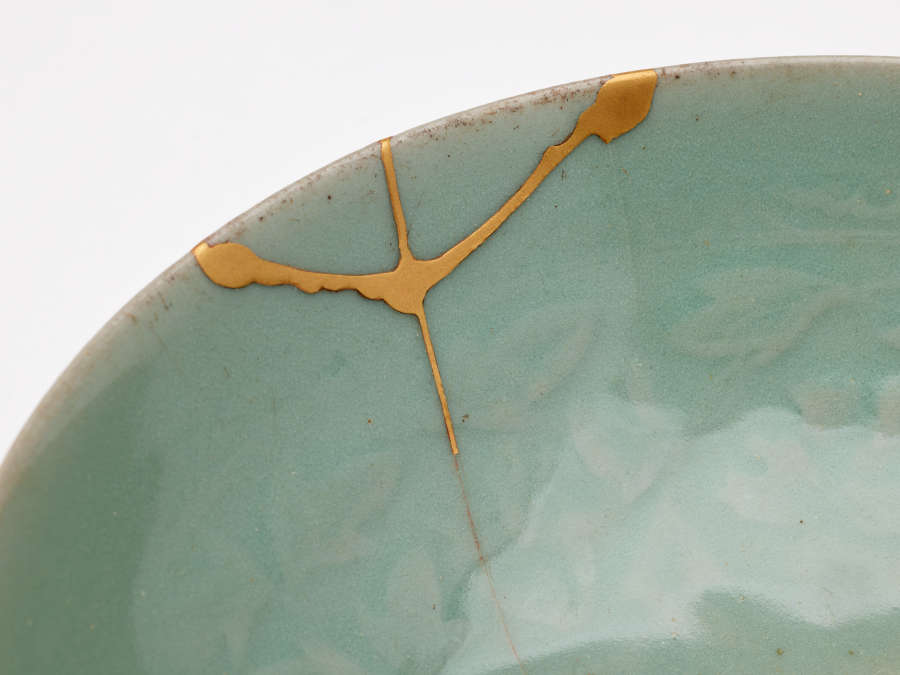 Detail of an edge of the inside of a teal bowl with cracked fragments patched together with golden cement that divide the faded floral pattern inside.