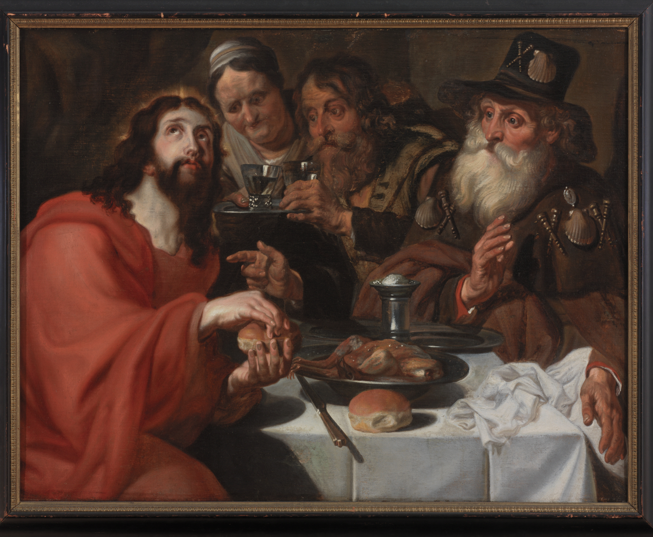 An oil painting depicting Jesus with a glowing halo and wearing a red robe seated at a table breaking bread with three other figures who are watching him intently.
