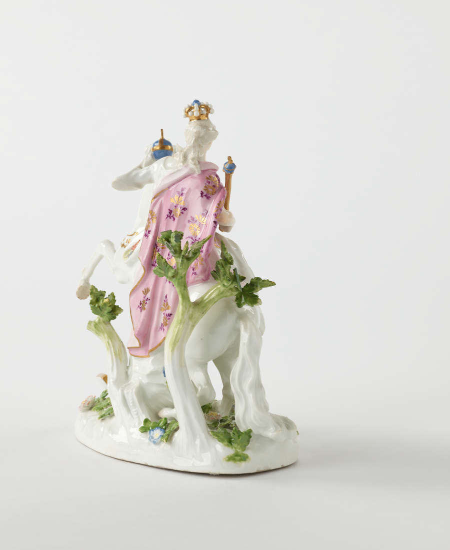 A sculptural figure on a rearing horse in historical clothing that is yellow, pink, and white with floral decorations. The figure is holding a staff and ball.