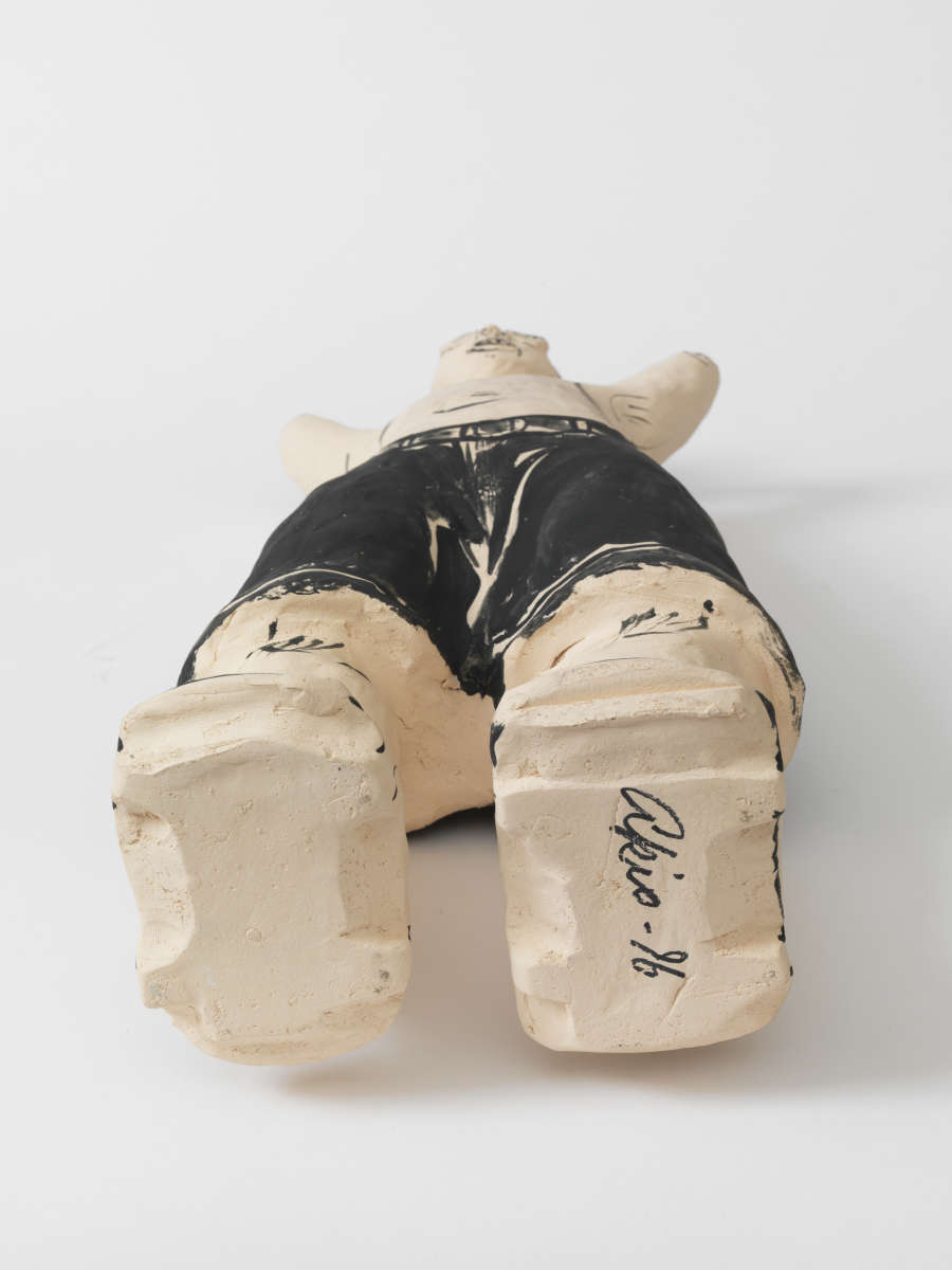 A ceramic figure lying on its back, shown from the feet upward. The unglazed soles reveal a thin black signature marking the foot to the right.