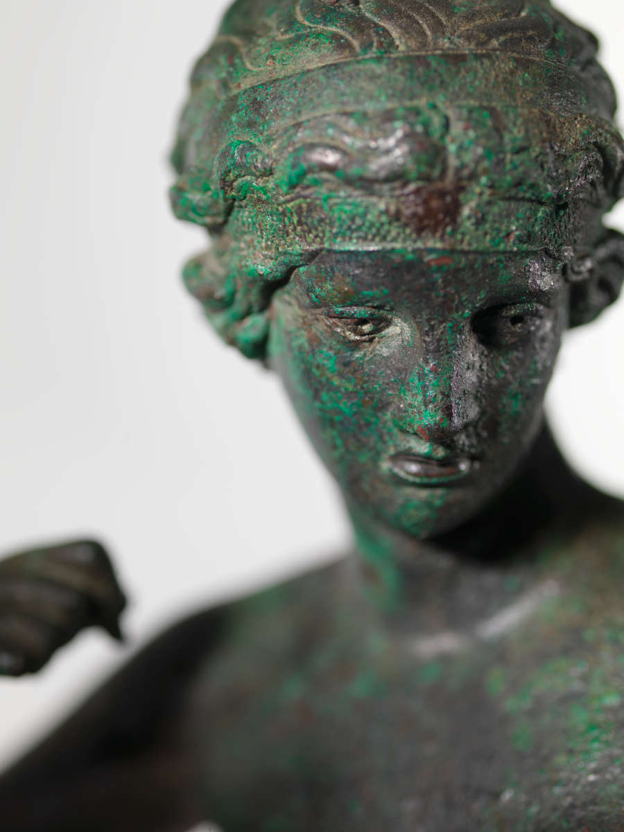 Detail view of a bronze statue of a crowned nude woman at an angle. Visible are the details of her face, hair and clenched fist curled towards her shoulder.