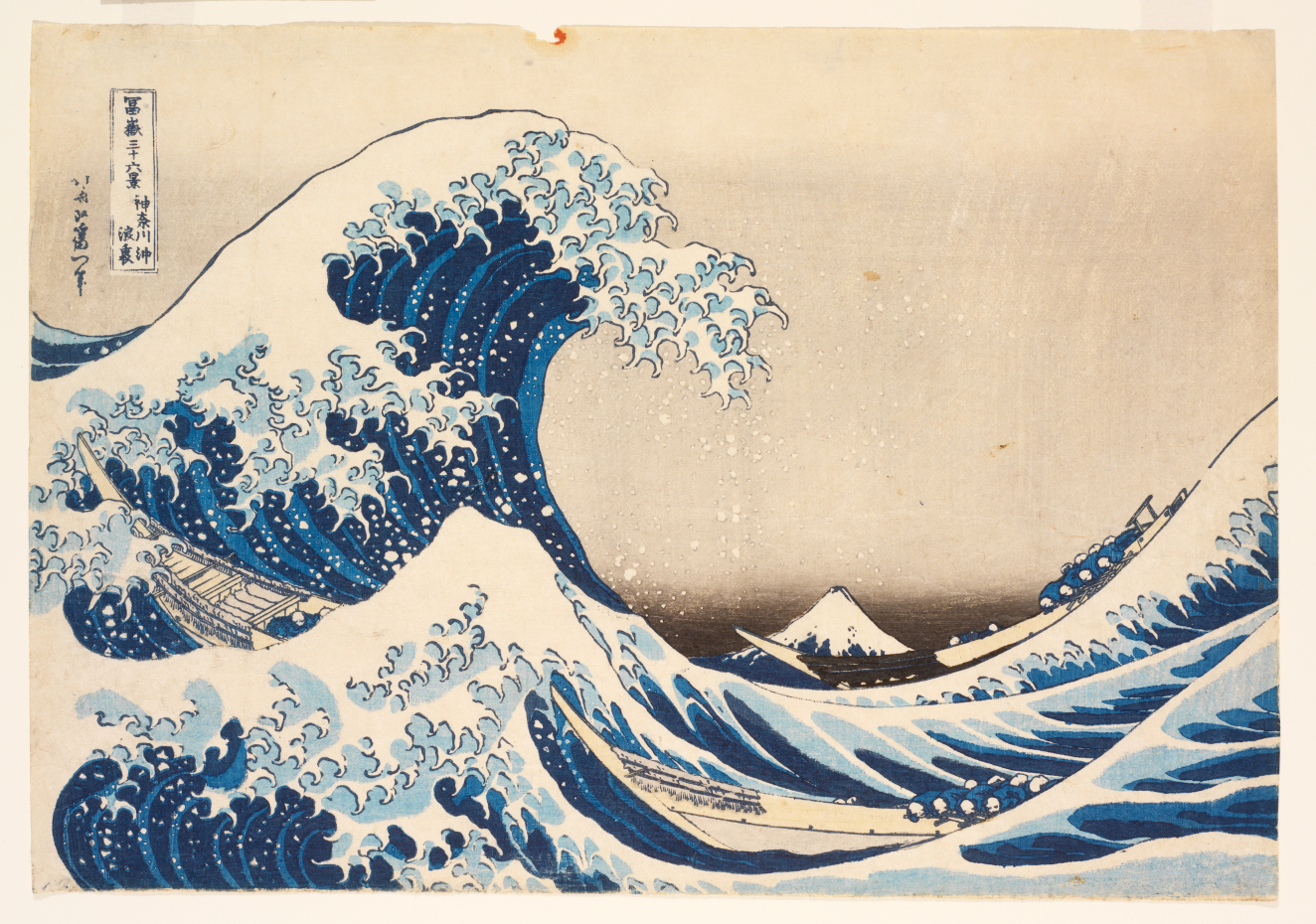 Woodblock print of a towering blue wave curling above two slender boats, spraying sea-foam and framing a distant Mount Fuji under a soft, stormy sky.