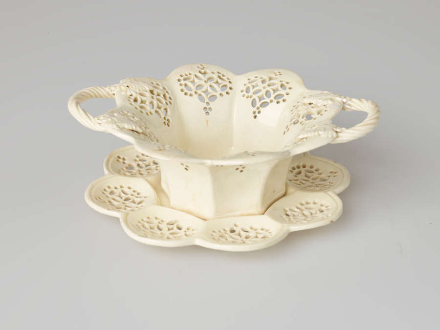 A cream-colored dish with two swirled handles and pierced decorations. Dish sits on a matching tray.