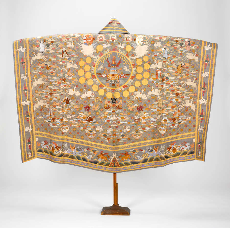 The intricate back of a priest’s robe hung on a pedestal, featuring a concentric arrangement of golden circles, pastel illustrations of fauna, monks, clouds and pagoda’s with illustrated borders.