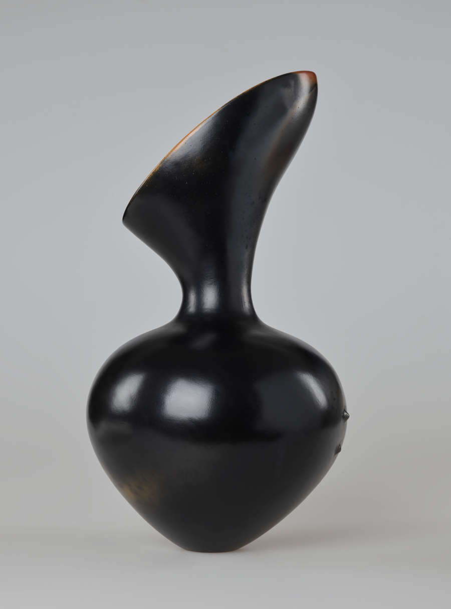An asymmetrical hourglass-shaped black vessel with a reflective finish. The top is slanted, opening diagonally to the left side. Two small bumps protrude from the right side of the base.