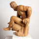 Realistic terracotta sculpture depicting a muscular bearded man facing away to the left, his bent arm supporting him while his leg sits over a vessel with a curved handle underneath.
