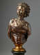 Bronze bust of a woman with regal expression, hair in twists, gold earrings, beaded necklaces adorning her collarbones, and a dark tightly-wrapped strapless top on a gold base.