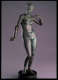 Gray metal sculpture with a green patina of a crowned nude woman standing on a circular pedestal, one arm down, grasping an object, and the other curled towards her shoulder. 