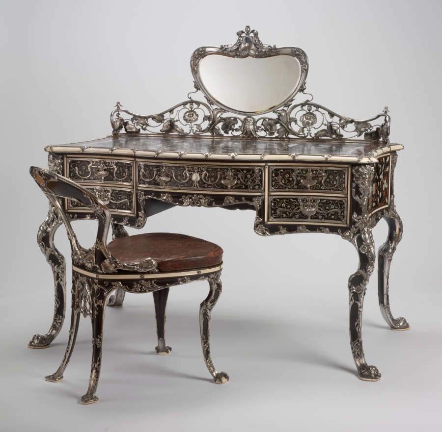 An ornate silver desk with a matching chair, both adorned with intricate floral inlay and curved legs. The desk features drawers and a central mirror framed in elaborate silverwork. 