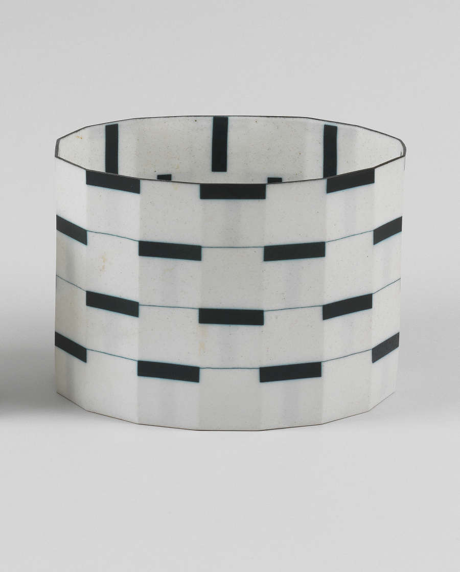 A cylindrical vessel constructed of vertical panels patterned with white squares interrupted by thin, staggered black bands, evoking piano tiles. 