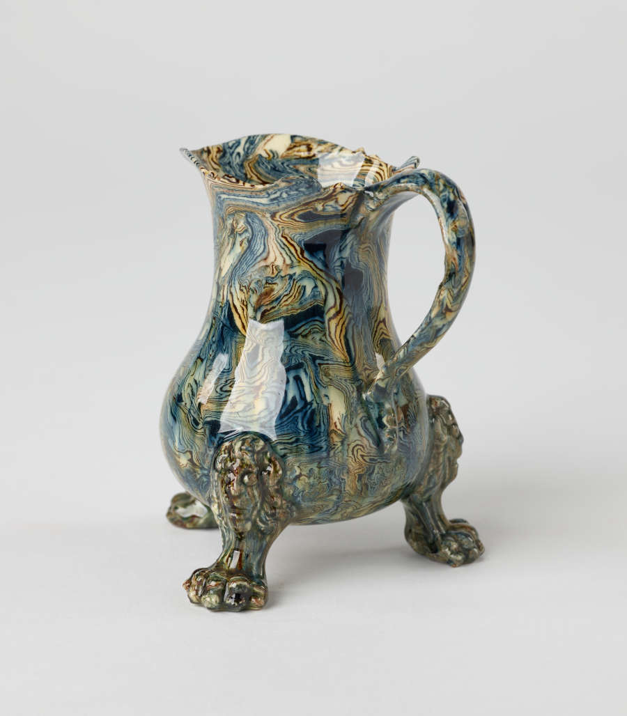 A small vessel with a cream, blue, and green mottled design. Three sculptural feet are shaped like lion’s paws with lion heads attached to body of vessel.