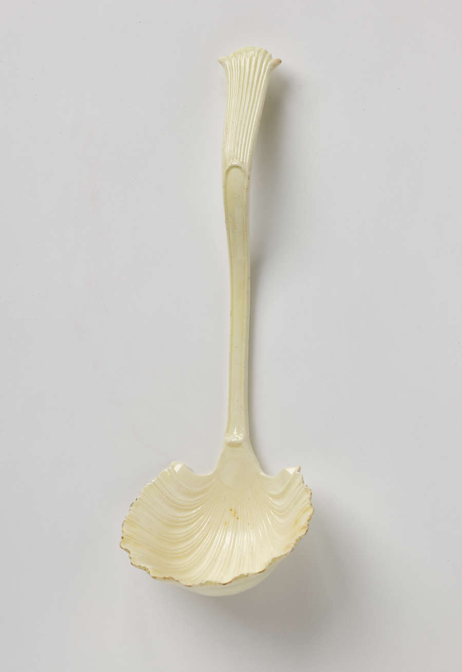 A cream-colored ladle with a shell shaped bowl and simple necked handle that comes to a flared out point with linear decorations.
