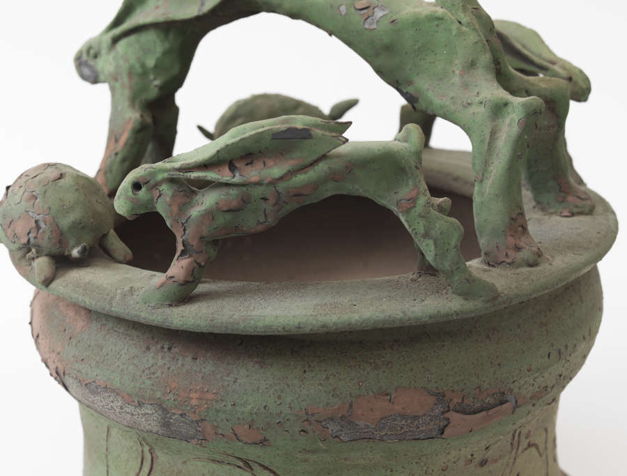 Close-up of a green ceramic vessel rim depicting a sculpted rabbit chasing behind a turtle along the edge, the surface mottled with reddish clay peeking through the green glaze.