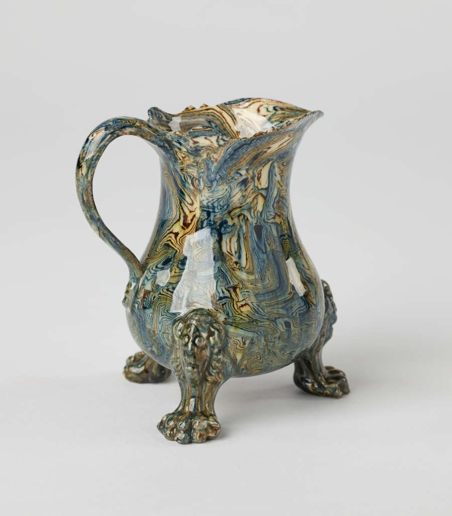 A small vessel with a cream, blue, and green mottled design. Three sculptural feet are shaped like lion’s paws with lion heads attached to body of vessel.