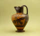 Tall onion-shaped jug with a pinched mouth forming a spout, and a handle. The black and orange jar is decorated with natural and geometric patterns and illustrations of a sphynx.