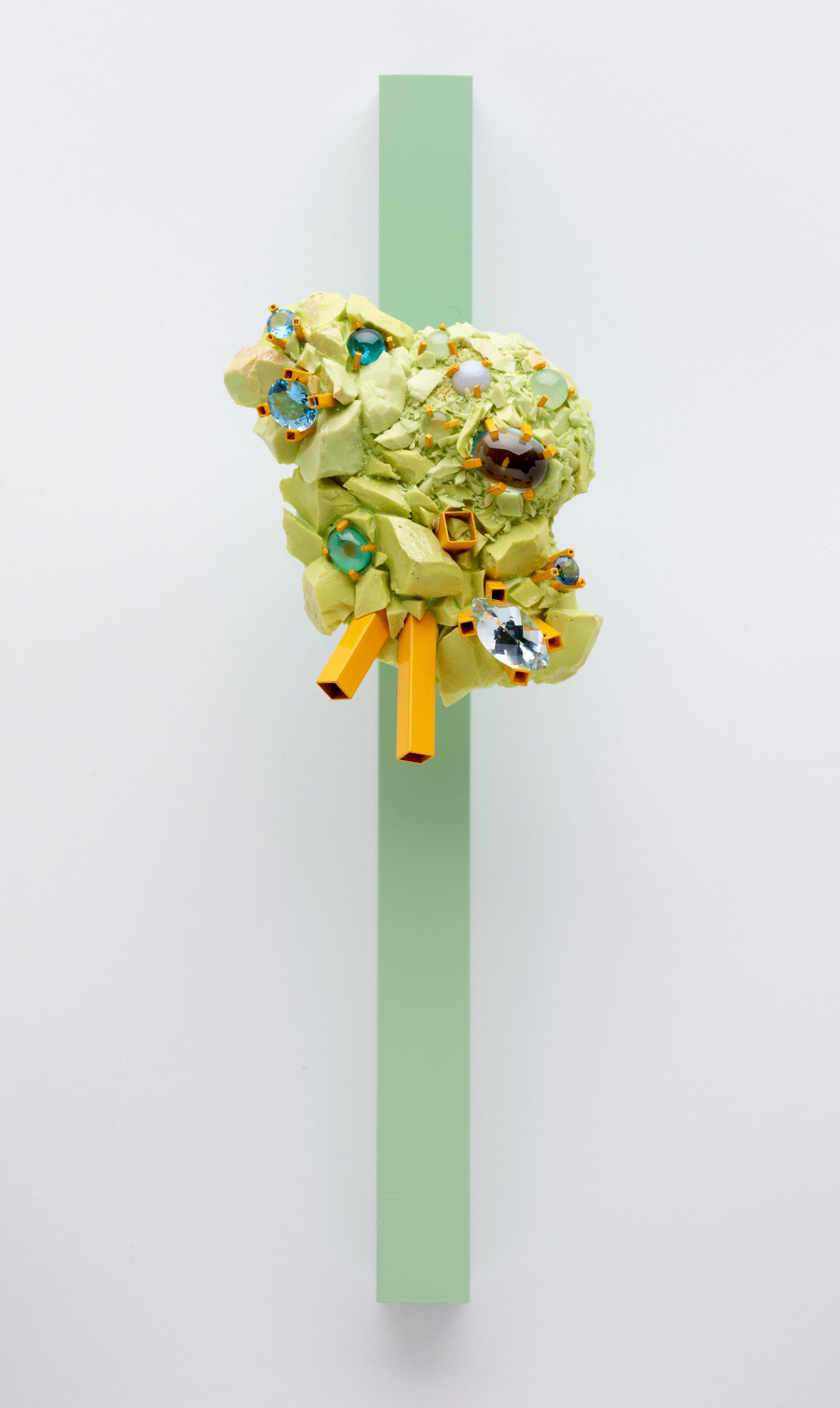 A sculptural piece of jewelry made up of a lime green cluster of shards and teal gems with vibrant orange accents, mounted on a slender, vertical, mint-green post.