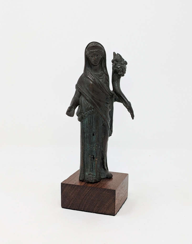 Dark greenish-black figurine wearing a large draped scarf and holding a horn-like object. With an outstretched hand, gestures towards the viewer and is placed on a rectangular dark brown platform.