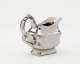  A silver creamer with a decorative handle and a rounded square body with sculptural decorations.
