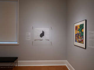 Image of installation view of Dominic Quagliozzi’s work: Artwork hangs on a gray wall with an object label on the left. Work features a dark gray abstract shape on white paper. Window and bench visible on the left. Another artist’s work is partly visible on the right. 
