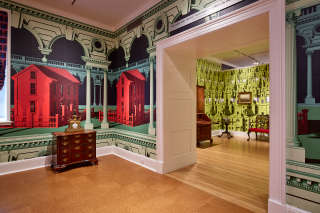Colorful wallpaper embellishes the walls of two adjoining rooms containing antique furniture. The foreground pattern includes large vibrant red houses and green architectural details. A chartreuse pattern is adjacent.