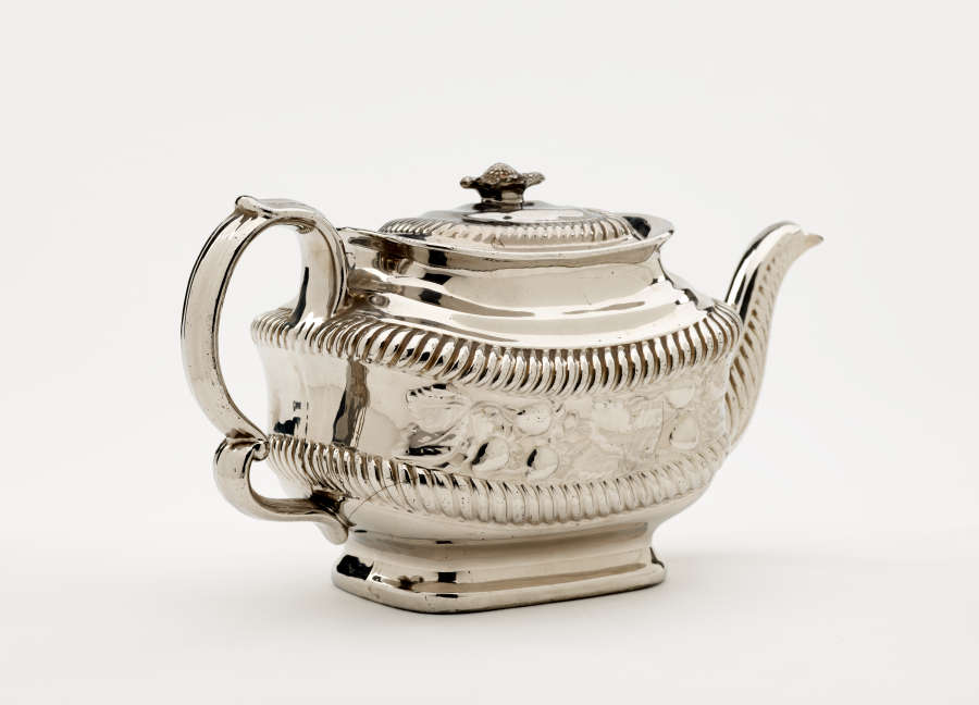 A silver teapot with a decorative handle, a rounded square body and spout with sculptural decorations.