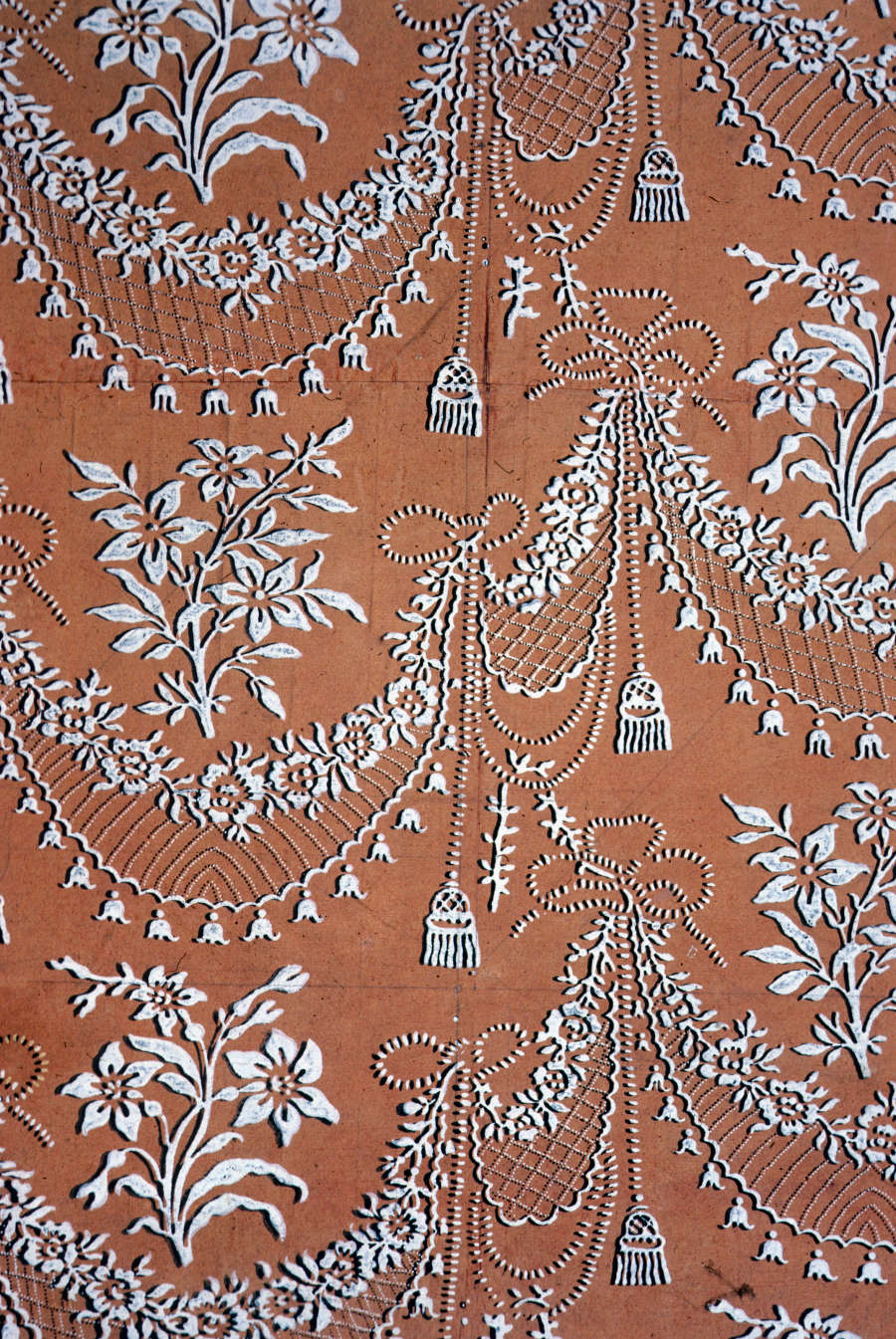 Intricate and elegant patterns of draping florals and tassel motifs done in white appear embossed on a warm dark orange backdrop