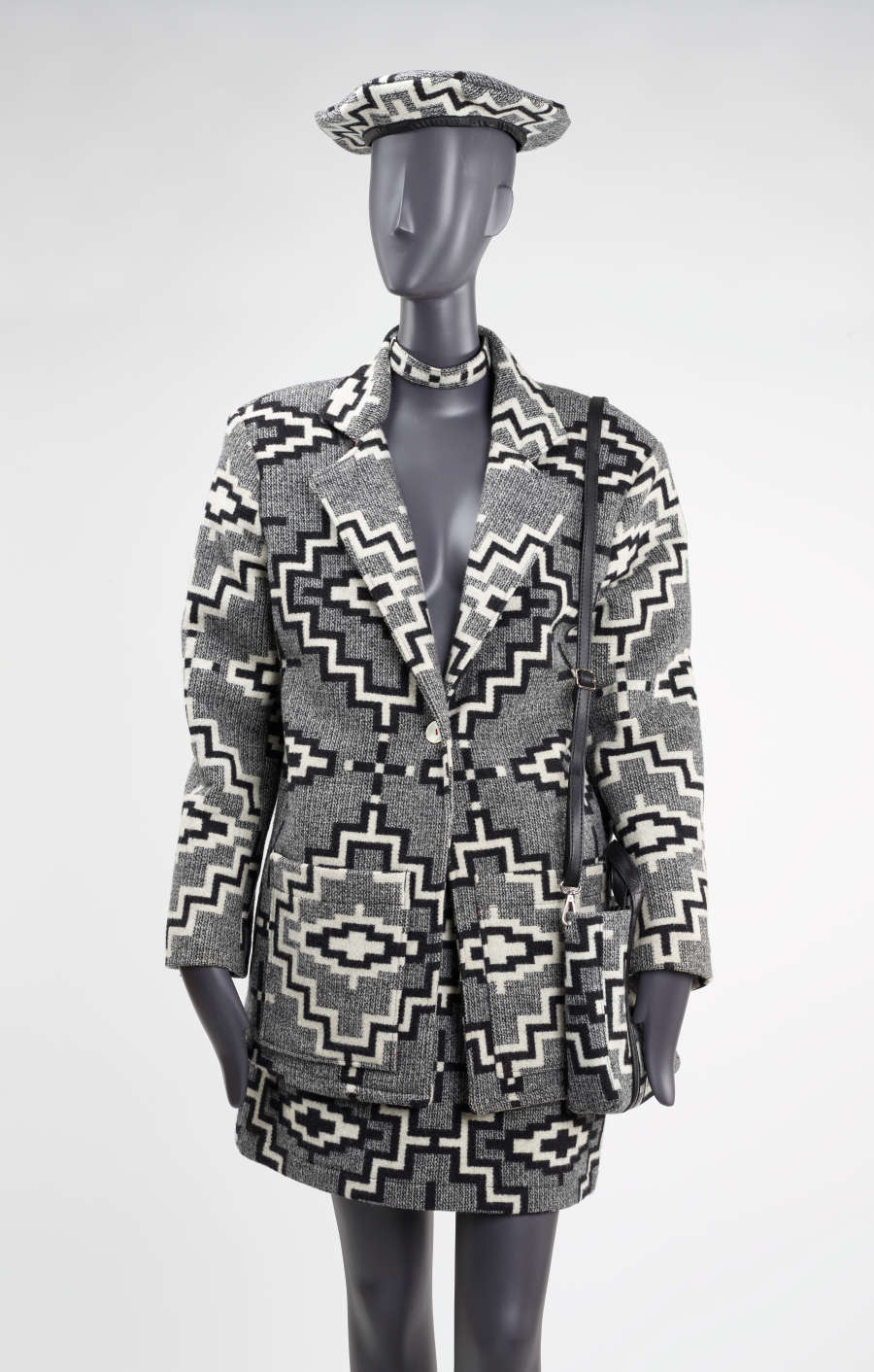 Mannequin dressed in a gray tweed black and white geometric patterned blazer with a matching hat, choker and bag. The skirt rests above the knee, and blazer reveals mannequin's chest.