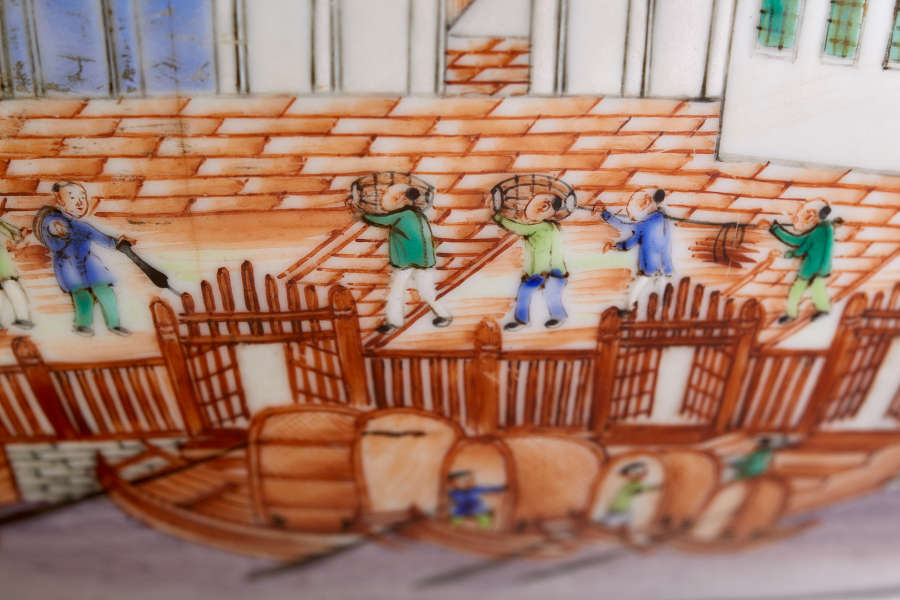 A close up of a painted ceramic depicting workers carrying baskets along an orange-tiled courtyard bordered by fences, with docked wooden boats in the foreground.