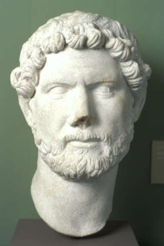 Marble bust from the neck up of a realistically carved bearded man with loose curly hair and furrowed brows resting on a pedestal against a gray backdrop.