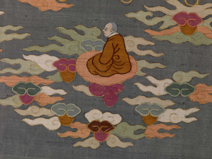 Section of illustrations of the blue robe’s back featuring a circular arrangement of wispy pastel clouds. A monk in ochre robes sits peacefully on a pastel red among them.