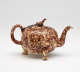 A mottled brown teapot with protruding feet, a spout, handle, and lid. The finial on the lid is connected to the handle by a chain.
