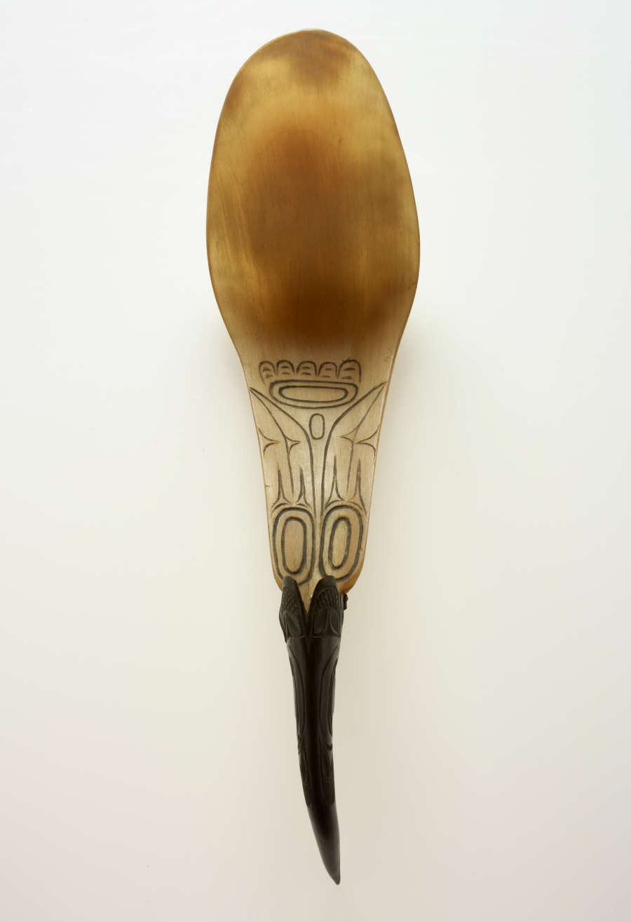 Top view of a ladle. The spoon’s bowl is round, extending into a narrower tan stem decorated with symmetric geometric motifs connecting to a pointed and thin black handle.