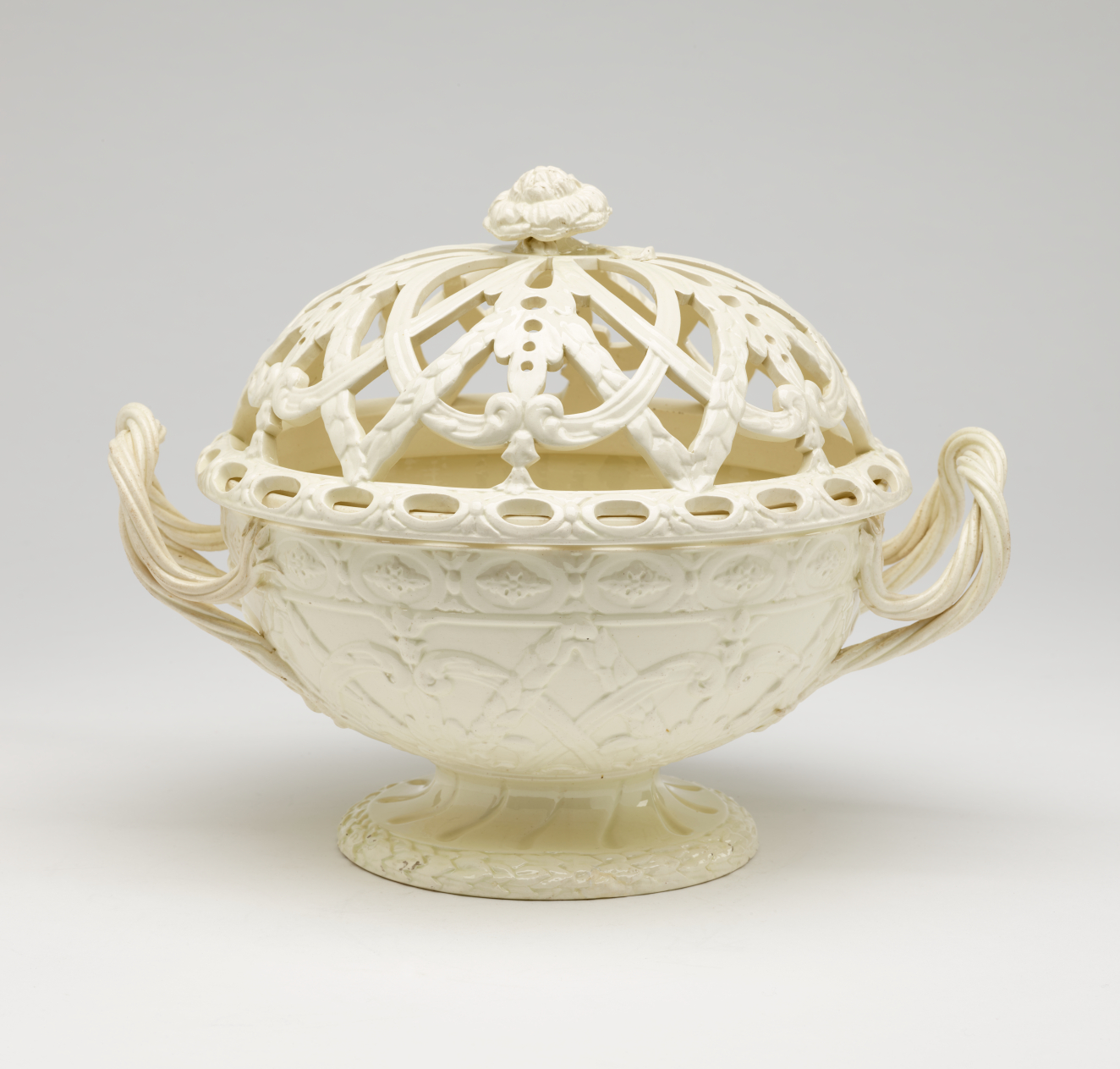 A cream-colored basket with sculptural woven handles, sculpted foot, and a lid with cutout designs.