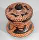 A lidded terracotta container with a black-glazed top, featuring a cracked lid adorned with black dogs and rays circling the handle. 