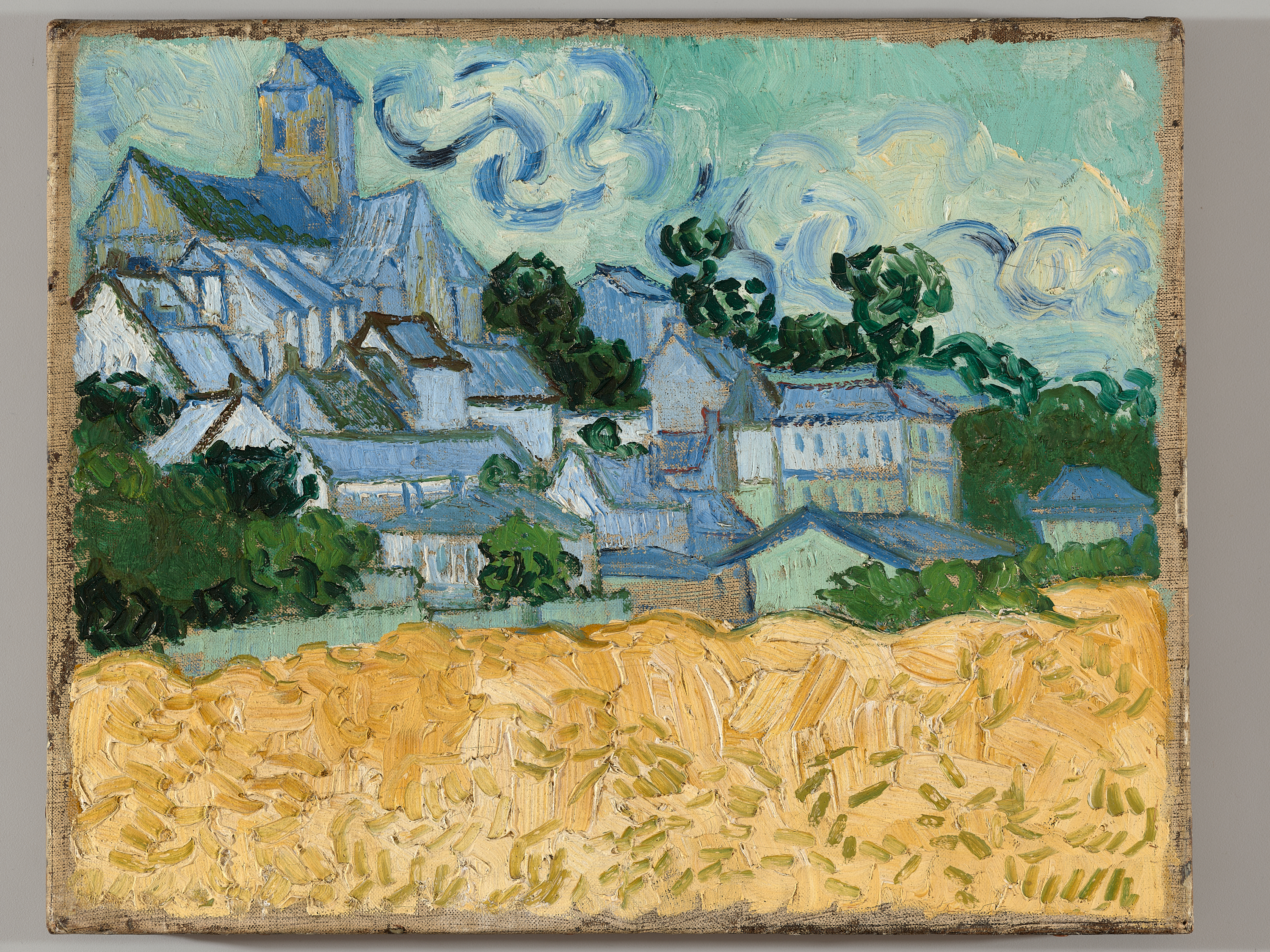 Landscape painting with thick, textured brushstrokes depicting an idyllic village scene of blue-gray rooftops rising among green trees above a golden field, the sky filled with swirling pale blue clouds.