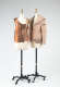Two dress forms with garments: a sleeveless top constructed of layered brown fabric and a beige, distressed jacket with exposed seams.