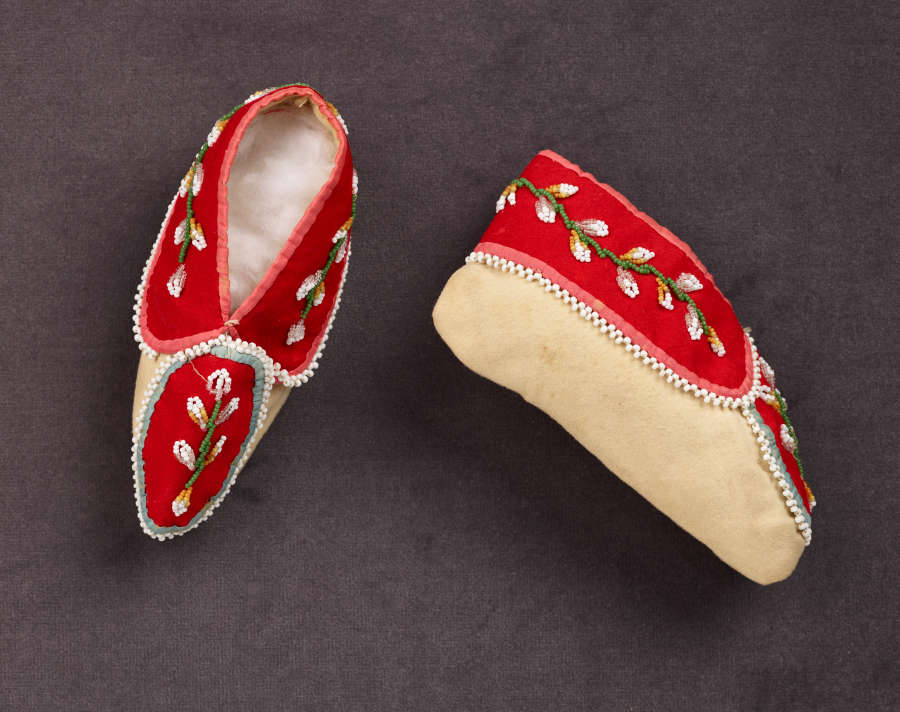 Pair of red moccasins featuring white and green floral embroidery as well as tan soles. One moccasin sits right-side-up on the gray surface while the other is on its side.