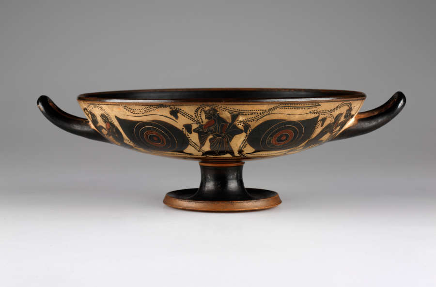 A wide, shallow, footed vessel with two handles on either side, curving upwards. The body is black and tan colored with an abstract design.