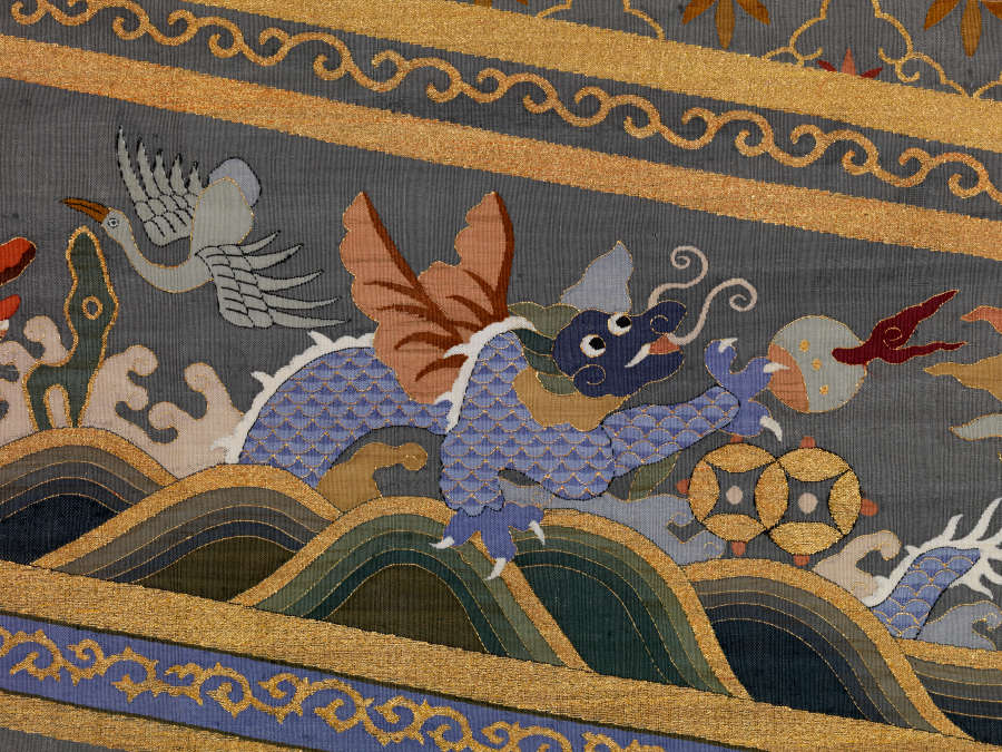 Border detail of the triangular tail of the robes back featuring a blue dragon in waves next to a soaring white bird against earthy-pastel clouds encased in golden striped borders.