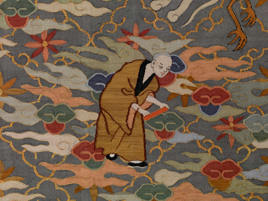 Robe’s back detail. A robed monk holds a red strip, amongst earthy pastel clouds, against a dark background with a thin golden diagonal grid, within each square is a floral motif.
