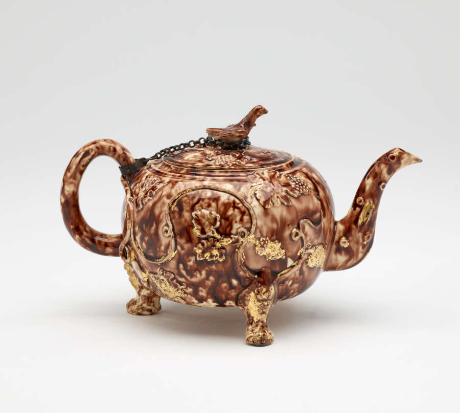 A mottled brown teapot with protruding feet, a spout, handle, and lid. The finial on the lid is connected to the handle by a chain.